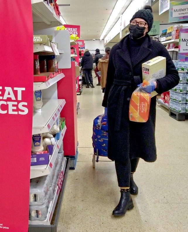 Kuvassa on ihminen kaupassa, katse tavarahyllyille kohdistettuna. Henkilöllä on maski ja kädessään kumihansikkaat sekä leipäpussi ja kainalossa maissihiutalepaketti.