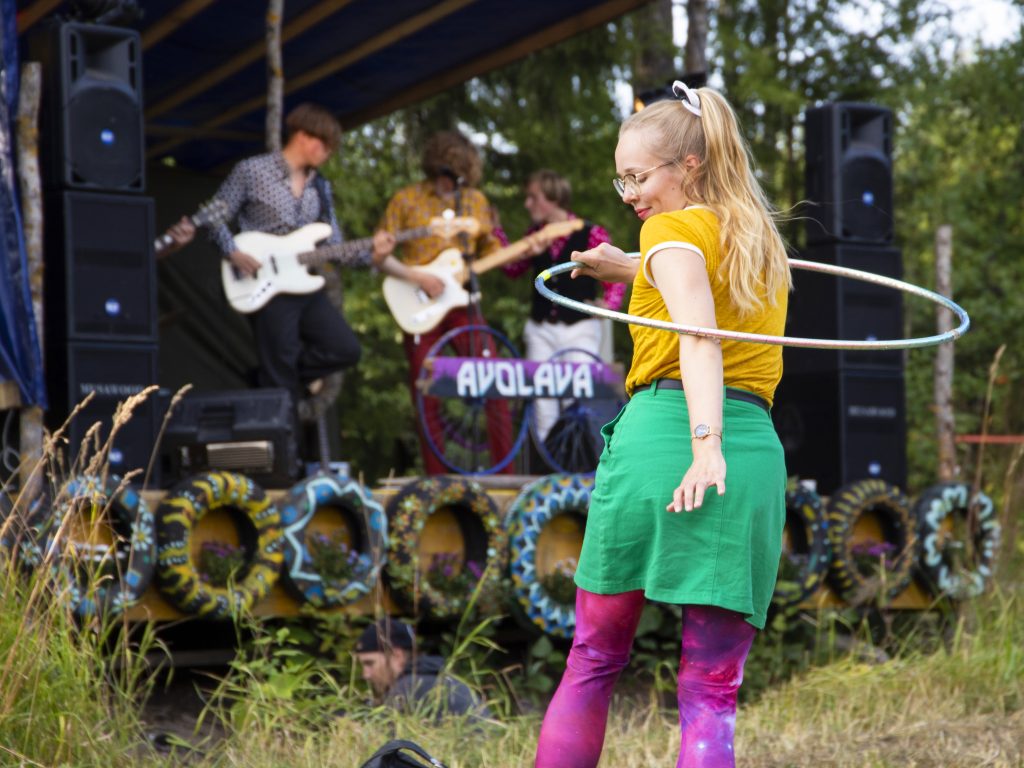 Kuvassa soittaa kaksi muusikkoa kitaraa lavalla, ja etualalla nuori nainen pyörittää hulavannetta ympärillään yllään kirkkaan väriset vaatteet.