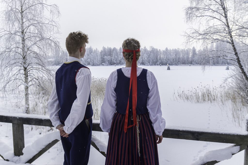 Kuvassa on kaksi kansallispukuista hahmoa selin katsojaan. He katselevat edessään olevaa talvista maisemaa.