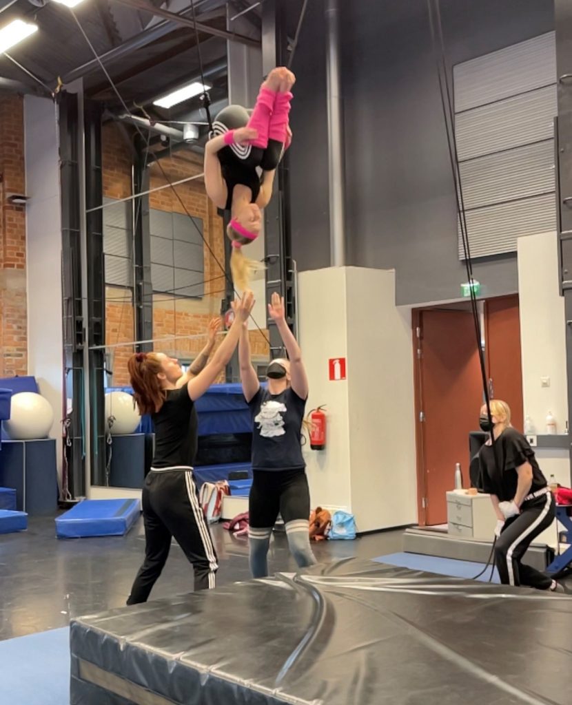 Kuvassa on neljä sirkusopiskelijaa harjoittelemassa akrobatiaa. Yksi heistä on ylhäällä pää alaspäin, ja kaksi hekilöä on hänen allaan kädet ylöspäin osoittaen, ottamassa kiinni, jos olisi tarpeen.