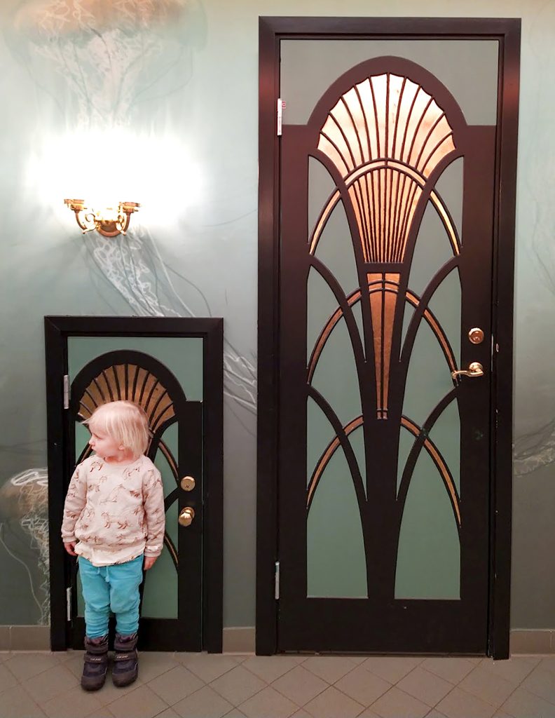 Kuvassa lapsiseisoo pienen, itsensä koruisen, oven edessä, ja vieressa on samanlainen ovi mutta korkeampi.