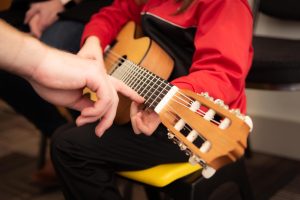 Kuvassa lapsi opettelee kitaransoittoa ja aikuisen käsi osoittaa otelaudan kohtaa.