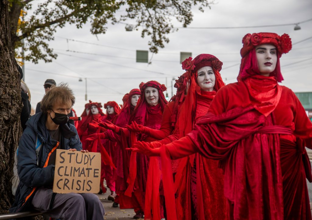 Kuvassa on punaisiin pukuihin pukeutuneiden ja valkoisella kasvonsa värjänneiden promootiokulkue, mielenosoituskulku. Kuvan vasemmassa laidassa istuu puun juurella henkilö, jolla on kasvoillaan musta maski ja kädessään pahvilappu, jossa lukee "study climate crisis".