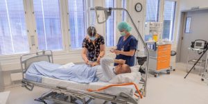 Kaksi opettajaa valmistelee hoitoluokassa potilasta esittävää henkilöä simulaatioharjoitukseen.