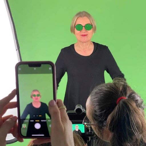 Kuvassa on Ilona Tanskanen vihreän greenscreening-kankaan edessä. Silmälasit on peitetty vihreällä kankaalla. Kuvan etualalla näkyy kännykän näytöllä sama näkymä, samoin kameran näytöllä.