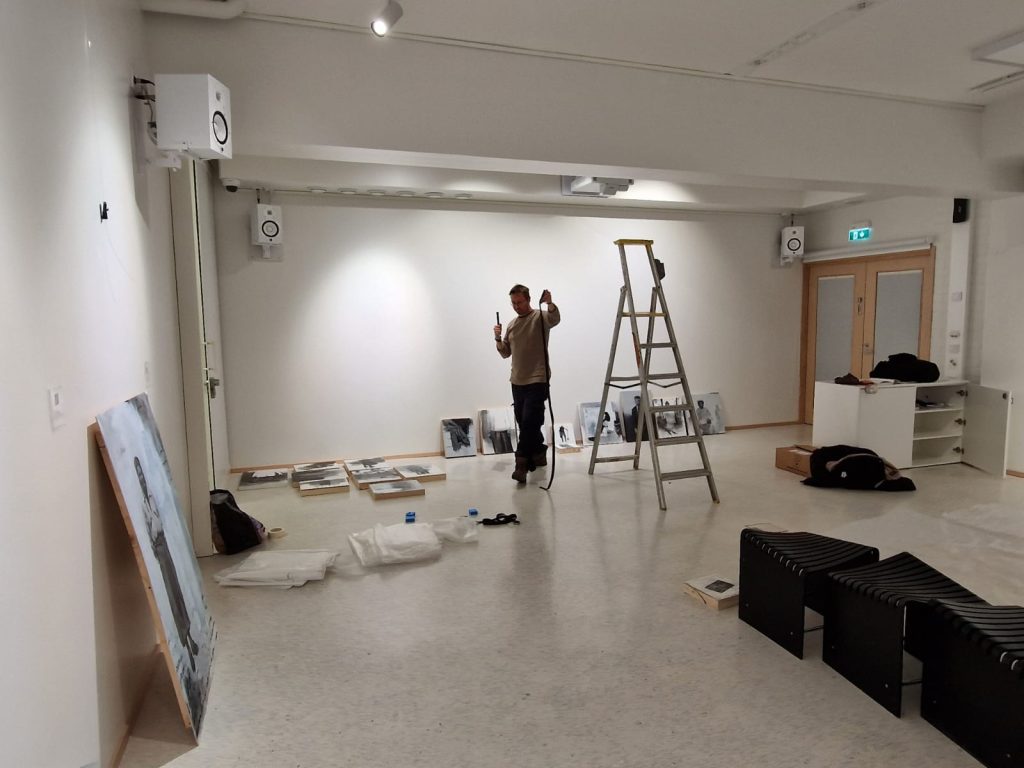Kuvassa näkyy taiteilija keskellä näyttelytilaa purkamassa näyttelyä. Tilassa on tikkaat ja kuvateoksia lattialla nojaamassa seinään.