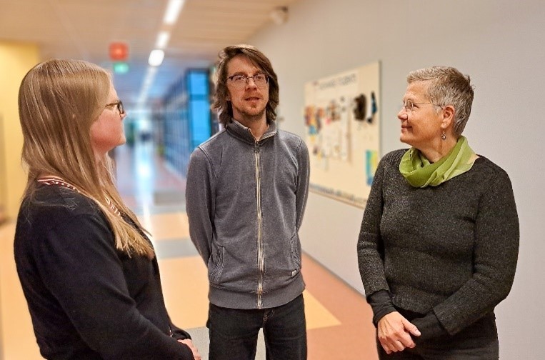 Kuvassa näkyy kolme henkilöä, joista kaksi reunimmaista katsoo keskellä olevaa, joka puolestaan katsoo suoraan kameraan.