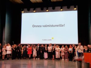 Kuvassa ovat Taideakatemian valmistuneet opiskelijat lavalla. Heidän yllään näytöllä lukee Onnea valmistuneille.