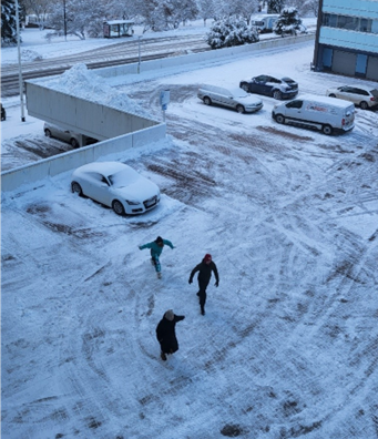 Kuvassa on talvinen näkymä kerrostalon pihalla ylhäältä päin kuvattuna. Keskellä on kolme henkilöä parkkipaikalla, jolla on myös autoja.