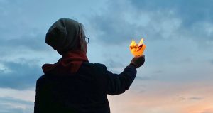 Kuvassa on henkilö selin kamaraan. Hänen kädessään on palavaa paperia. Taustana on hämärtyvä taivas.
