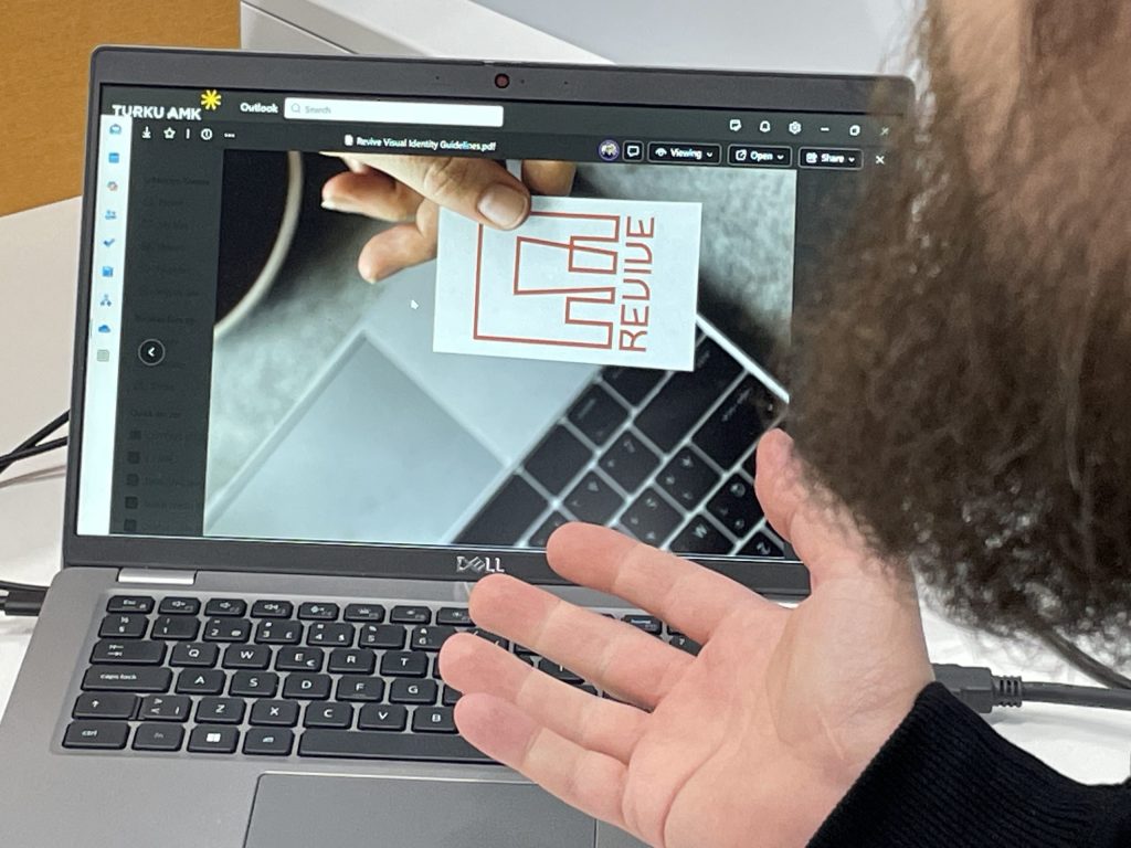 Kuvassa näkyy läppäri, jonka ruudulla on kuvakortti, jossa lukee Revive. Kuvassa näkyy myös avoin kämmen läppärin näppäimistön yllä.