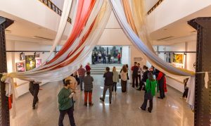 Kuvassa on galleriatila ylhäältä alaspäin kuvattuna. Ihmiset kiertelevät tilassa, jonka seinillä on teoksia.