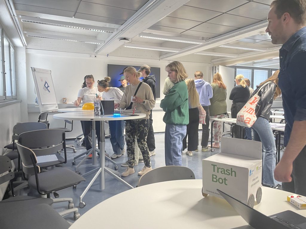 Kuvassa on ihmisiä luokkahuoneessa Taideakatemiassa. Etualalla on pyöreitä pöytiä, joiden äärellä seisoo ihmisiä.