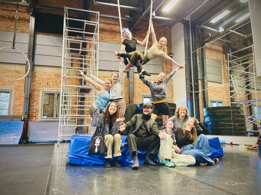 Kuvassa on ihmisryhmä sirkuskoulutuksen tiloissa. Lattialla paksun patjan päällä istuu viisi nauravaa henkilöä, ja heidän takanaan seisoo kolme iloisen oloista henkilöä kädet ylös ojennettuina. Heidän yllään on kaksi henkilöä narujen varassa niin ikään iloiset ilmeet kasvoillaan.