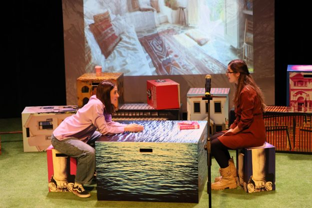 Kaksi naista näyttämöllä istumassa laatikoilla ja välissään pöydän kaltainen laatikko.