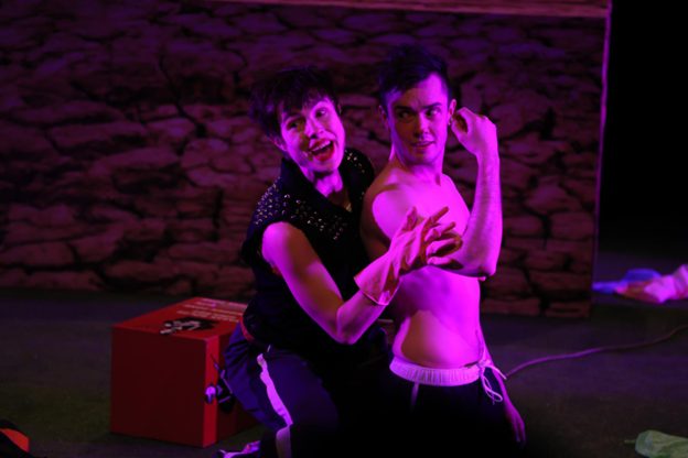 Kaksi esityksen henkilöä näyttämöllä lilassa valossa.