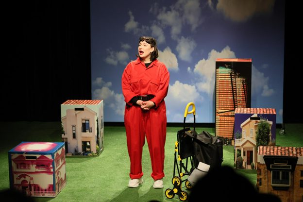 Esiintyjä näyttämöllä punaisessa haalarissaan ja punattuine huulineen laatikoista muodostuvien lavasteiden keskellä taustanaan poutapilvisen sinitaivaan kuva.