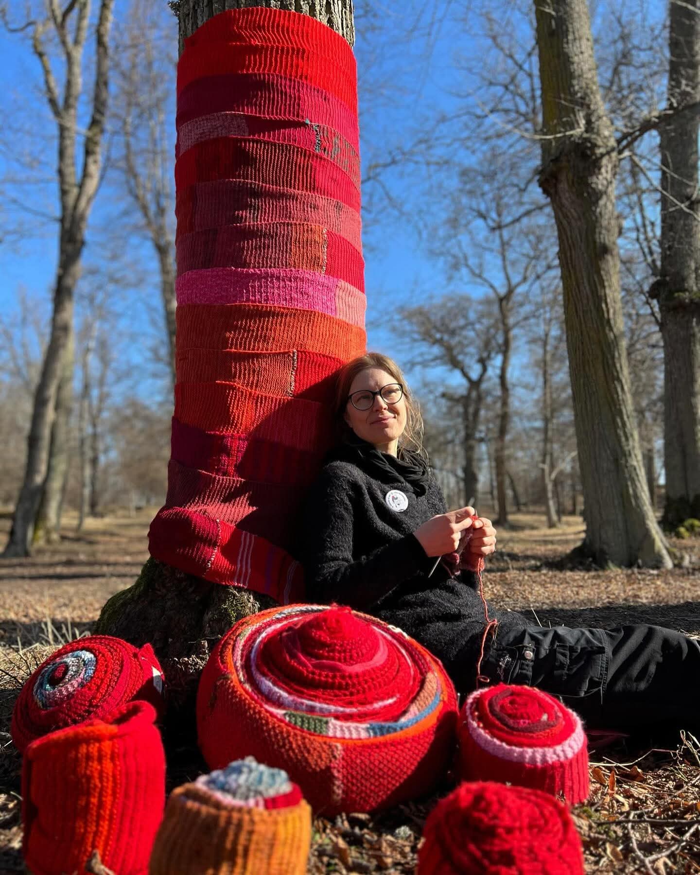 Nainen istuu, neuloo ja nojaa neulehuivilla peitettyyn puuhun. Edessä neulekäsitöitä.