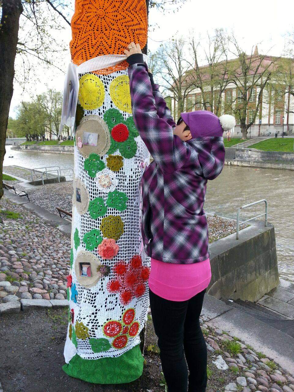 Kuvassa henkilö kiinnittää puuhun käsityötä. Taustalla joki ja sen toisella puolella talo.