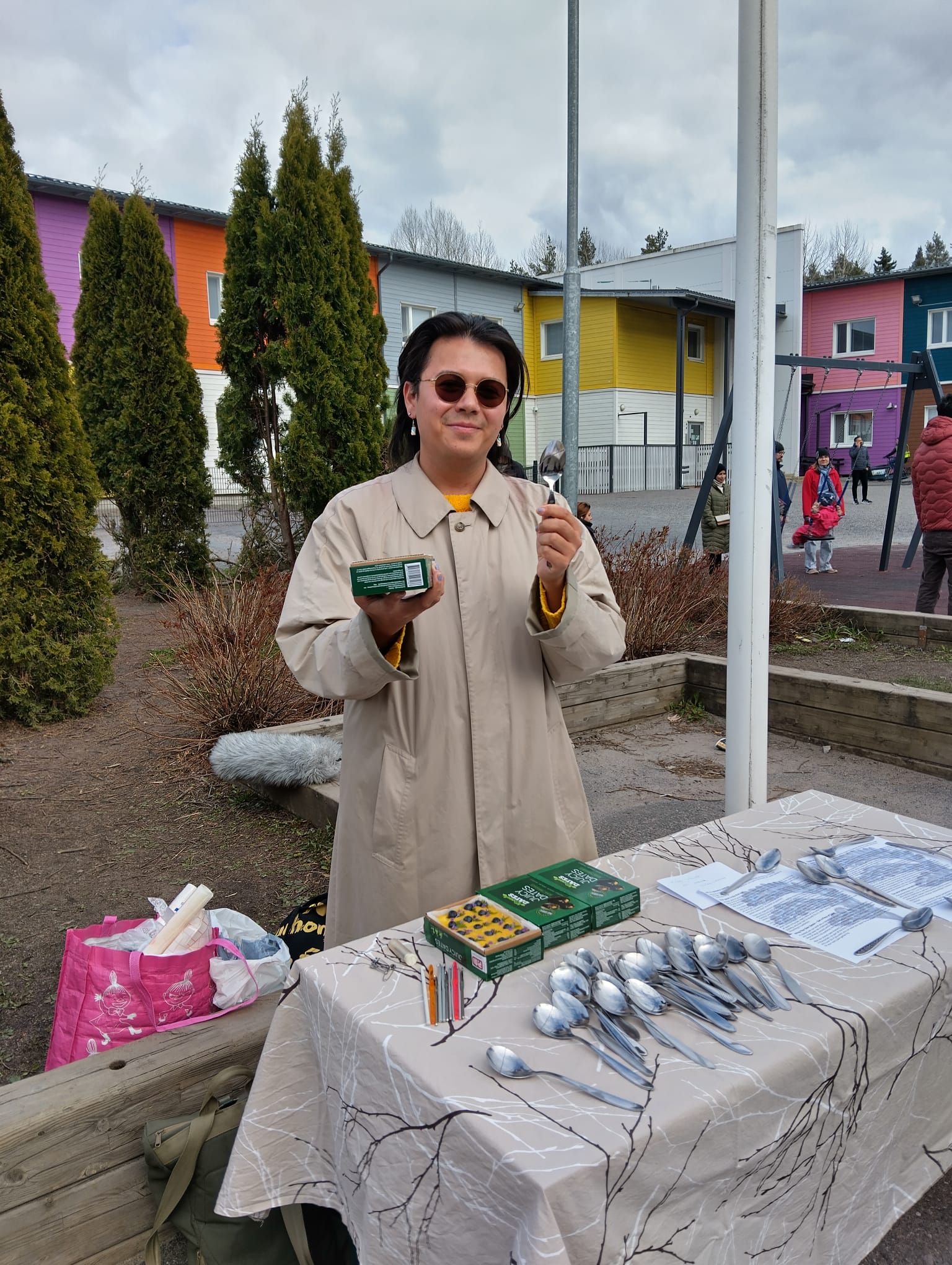 Aurinkolasipäinen henkilö pitää lusikkaa ja purkkia kädessään, taustalla puita, taloja ja lapsia leikkikentällä.