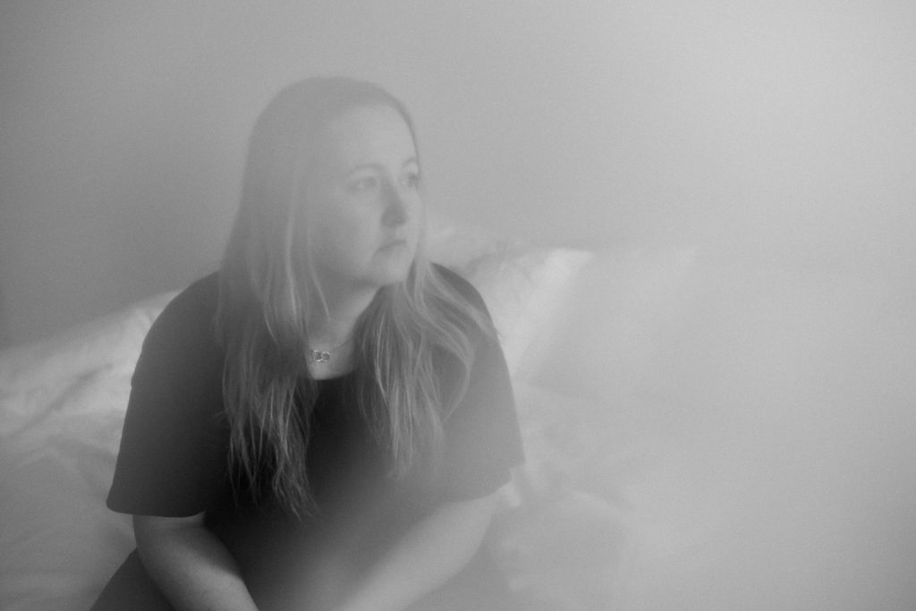 Kuvassa naisoletettu katselee oikealle istuessaan sängyllä.