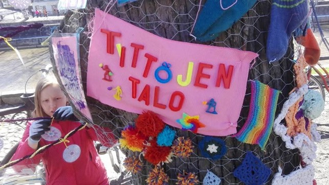 Tyttö kiinnittää puuhun kiinnitettyyn naruun kuvaa, etualalla paperilla teksti tyttöjen talo.