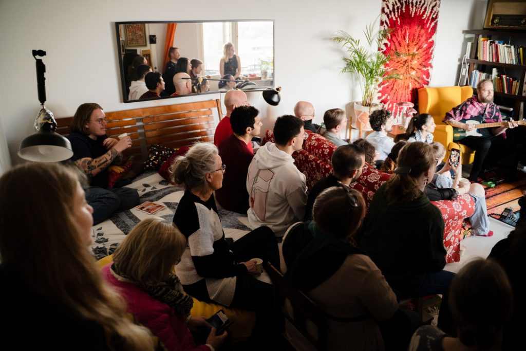 Kuvassa näkyy yleisö istumassa yksityiskodin olohuoneessa kuuntelemassa musiikkia.