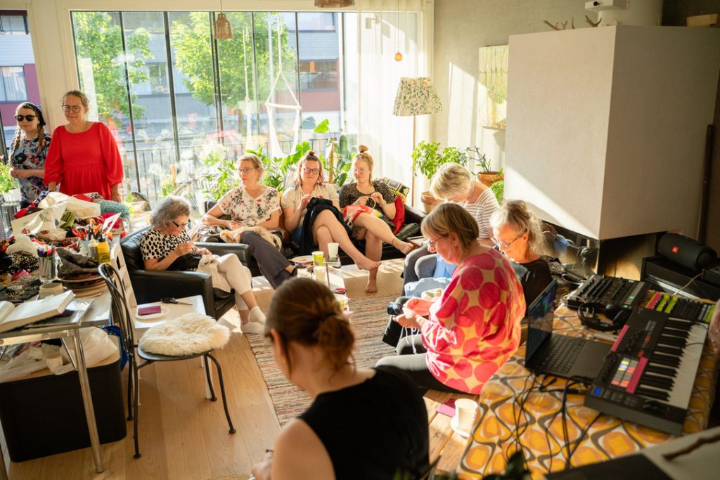 Kuvassa on ihmisiä aurinkoisessa olohuoneessa tekemässä käsitöitä. Kuvan oikeassa reunassa on koskettimisto, tietokone ja äänentoistolaite.