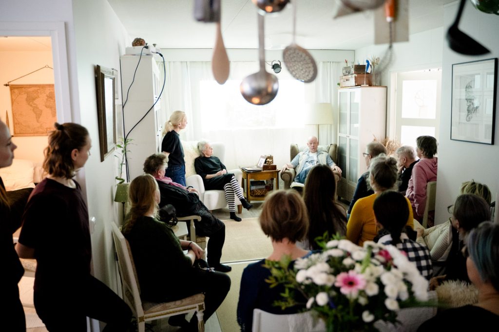 Kuvaaja on yleisön takana ja näyttää yleisön olohuoneessa seuraamassa ikkunan edessä olevilla nojatuoleilla istuvien keskustelua.