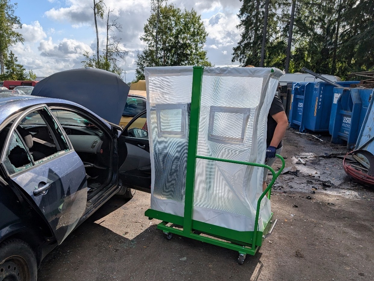 Pihalla auton ovesta poistettavan ikkunan alle on asennettu lasimurskeen keräämiseen tarkoitettu kärry. Taustalla puita ja pilvinen taivas.