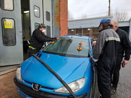 Kuvassa kolme henkilä on irrottamassa sinisen auton laseja pihamaalla.