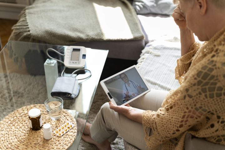 Ikääntynyt naishenkilö istuu sohvalla. Kädessään hänellä on tabletti, jolla on videoyhteys lääkäriin. Sohvan edessä pöydällä on lääkkeitä ja verenpainemittari.