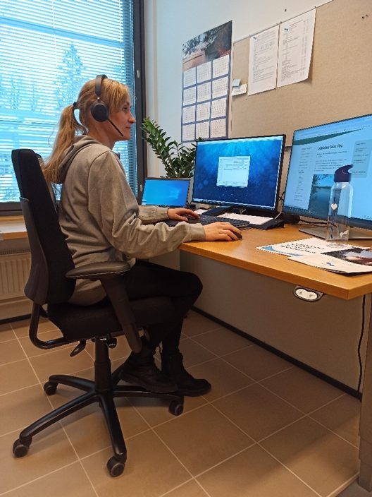 Toimistotyöntekijä työskentelee työpisteellään usean näytön äärellä kuulokkeet päässä, tietokoneen ja asiakirjojen parissa.